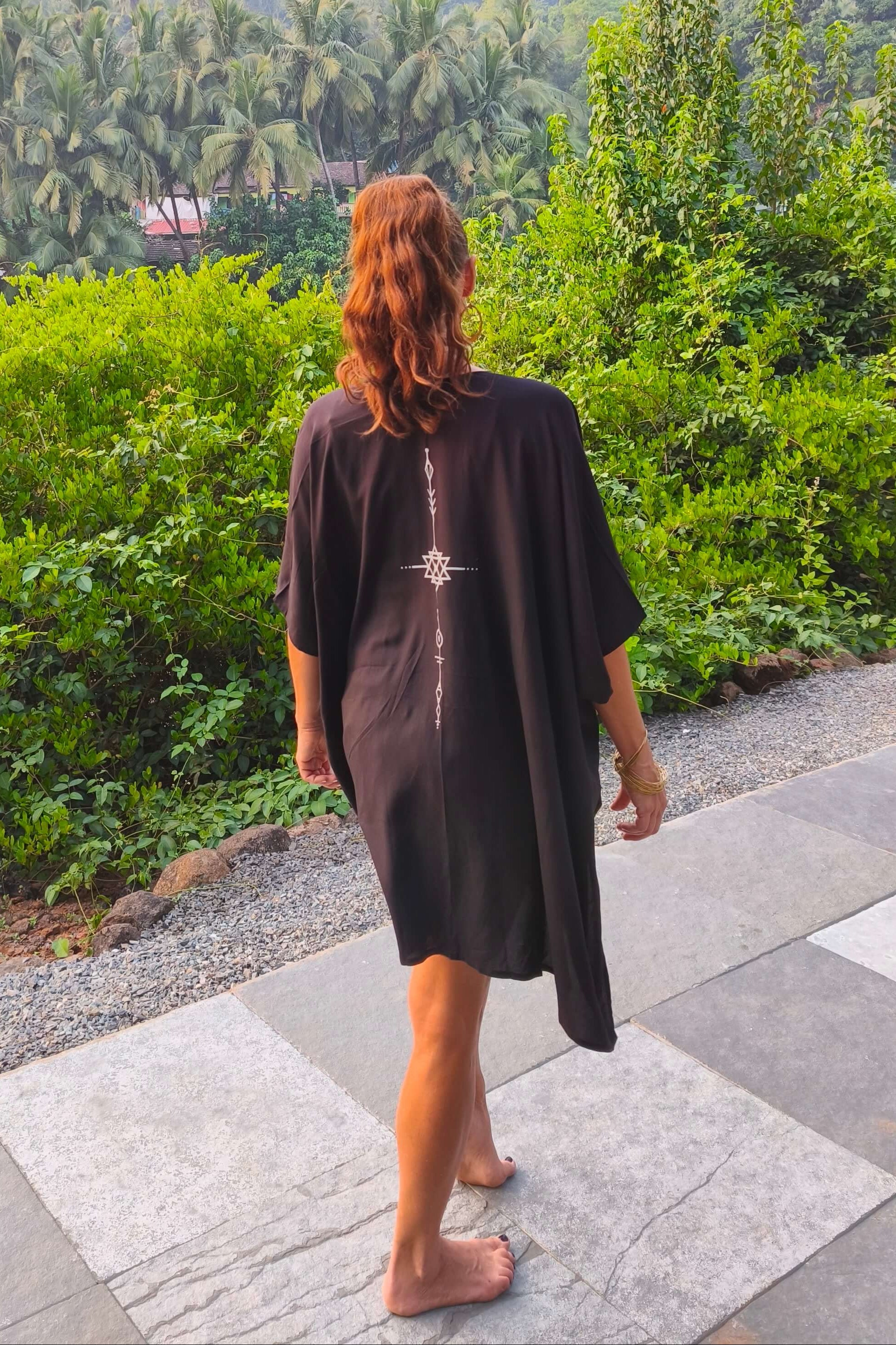 Person wearing a black kimono with a white symbol print on the back, standing on a stone path with greenery in the background.