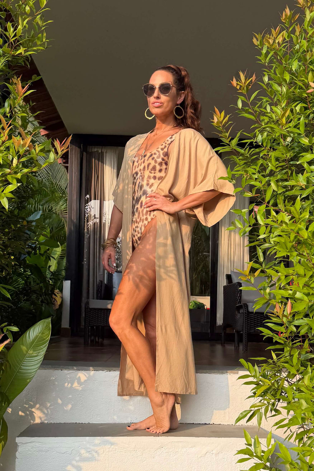 Woman in a beige long kimono standing on a set of white steps surrounded by greenery.
