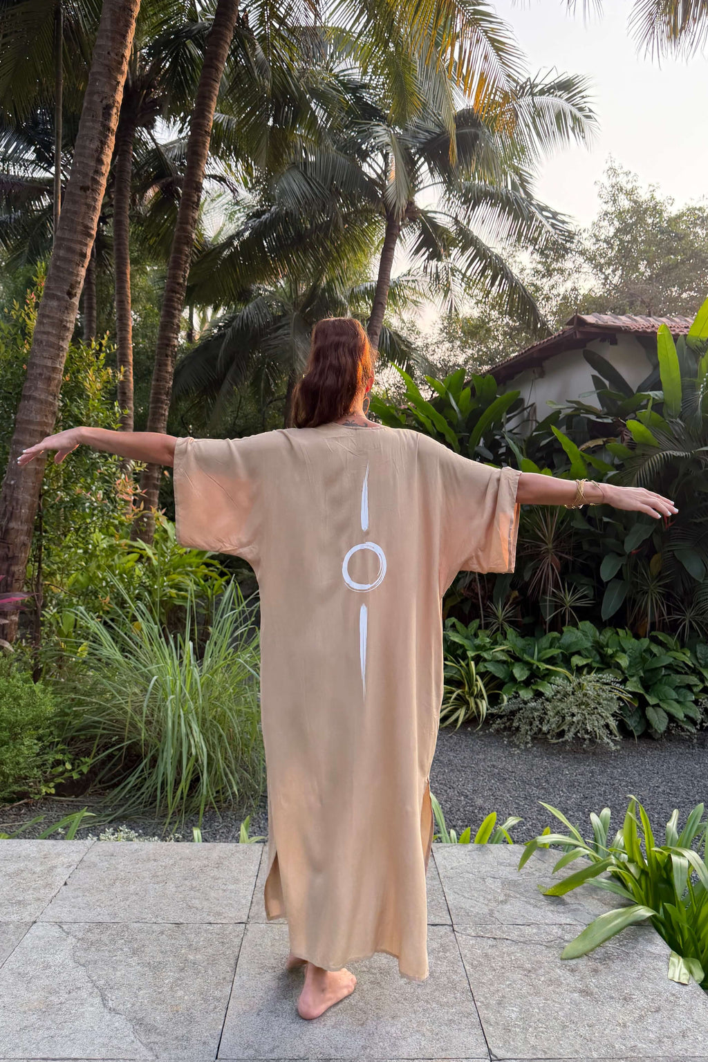 Person wearing a sand long kimono with a symbol, standing in a tropical garden.