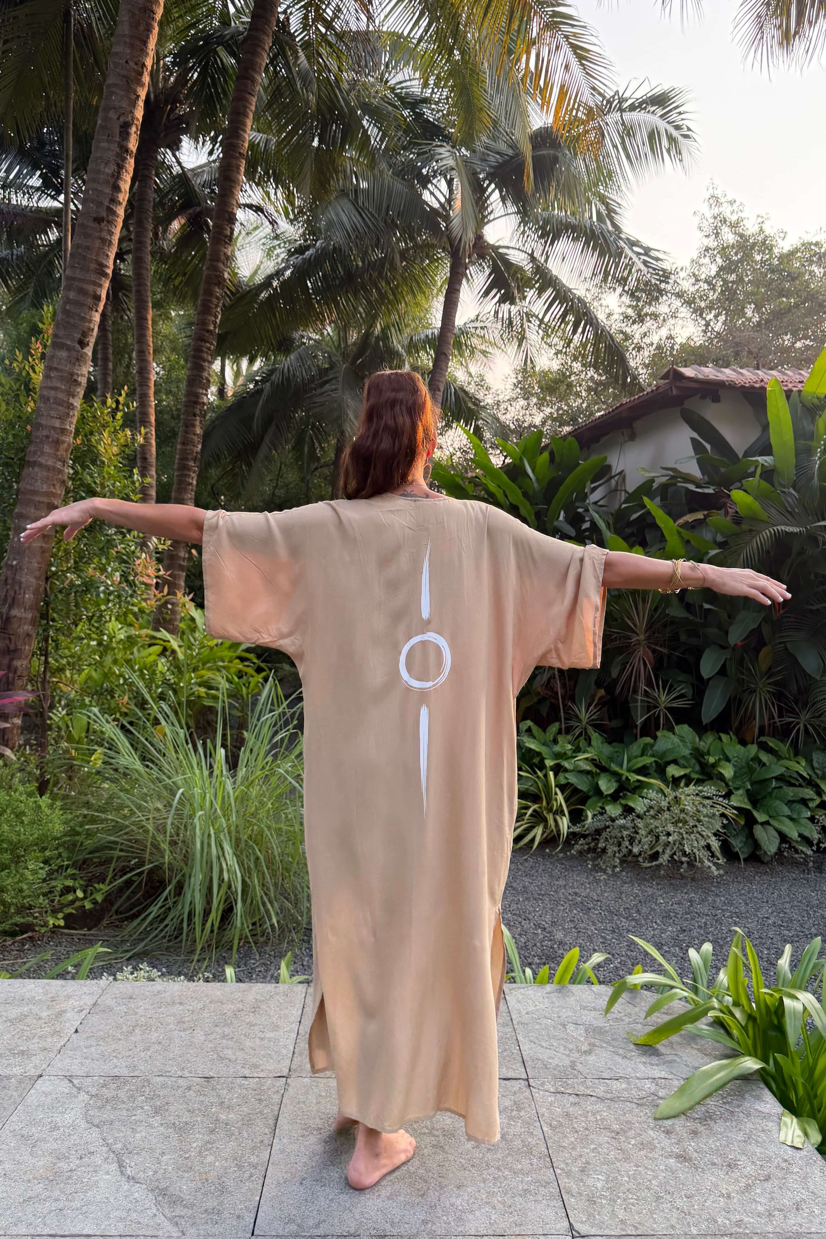 Person wearing a sand long kimono with a symbol, standing in a tropical garden.