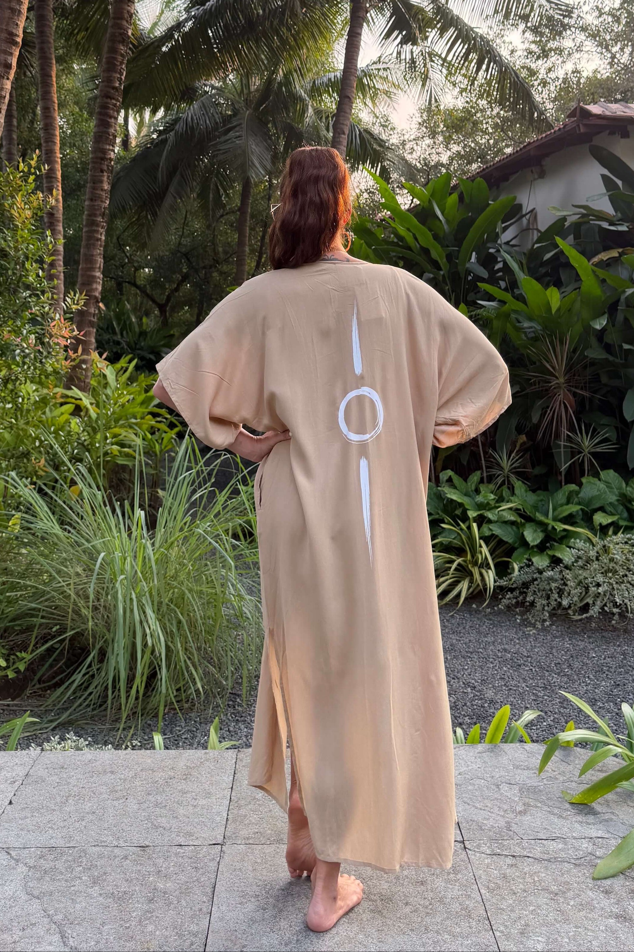 Person wearing a beige long kimono with symbol printed on it, standing in a tropical garden.