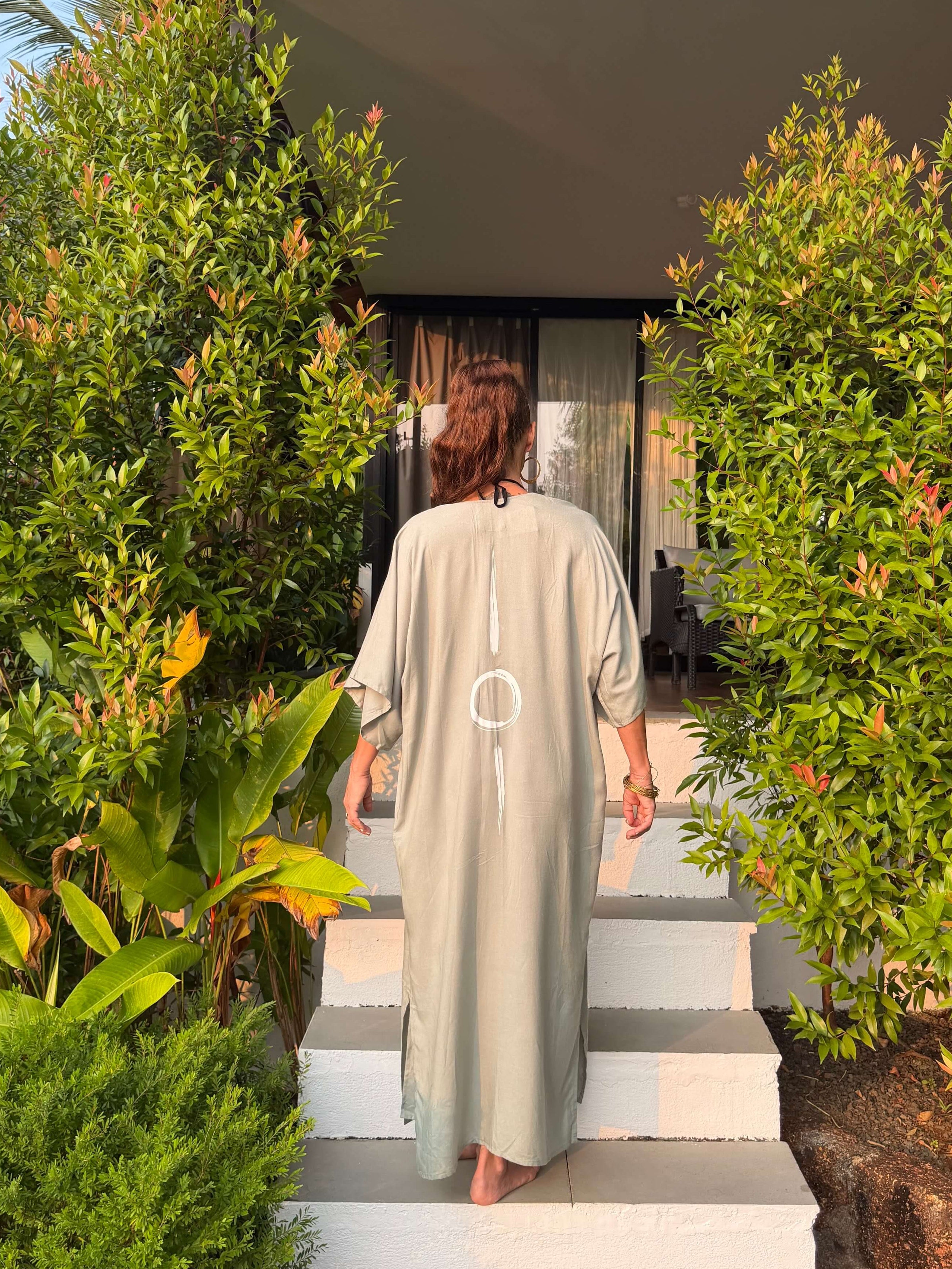 Person in a long light-colored kimono walking up steps towards a building with greenery around.
